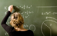 Teacher in a long black sleeve shirt with a blonde bun writing equations on a green chalkboard