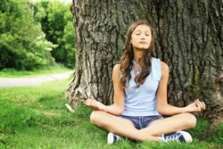Girl in a blue tank top and shorts sitting cross legged in front of a tree with her eyes closed