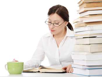 Woman with a dark ponytail and glasses wearing a white button up and reading next to a mug and a stack of books