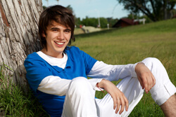 Boy in a blue polo and white pants sitting against a tree and smiling