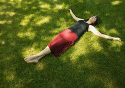 Woman in a black tan-top and long maroon skirt lying on the grass, with arms outstretched and eyes closed